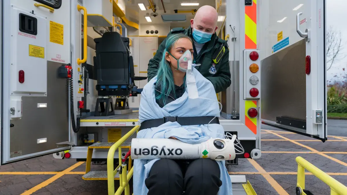 Two students are in front of an ambulance, one is dressed in Paramedic uniform, the other is wrapped in a blanket with an oxygen mask