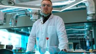 A man with piercings and tattoos is wearing a white lab coat and poses in a laboratory
