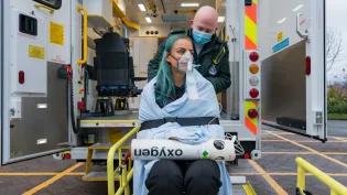 Two students are in front of an ambulance, one is dressed in Paramedic uniform, the other is wrapped in a blanket with an oxygen mask