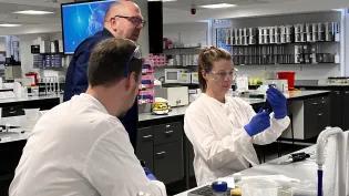 two students in white lab coats are helped by a lecturer 