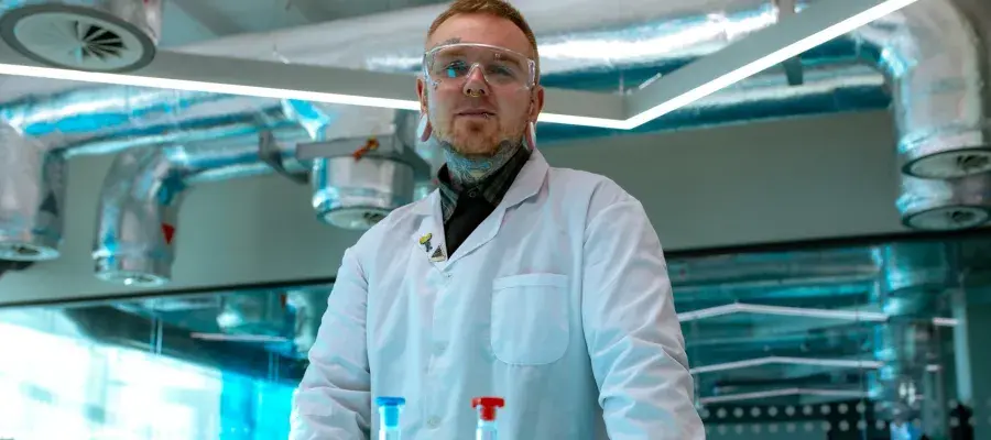 A man with tattoos and ear plugs poses in a white lab coat in a laboratory