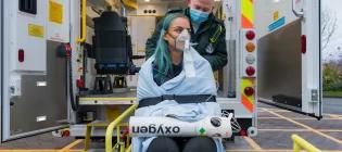 Two students are in front of an ambulance, one is dressed in Paramedic uniform, the other is wrapped in a blanket with an oxygen mask