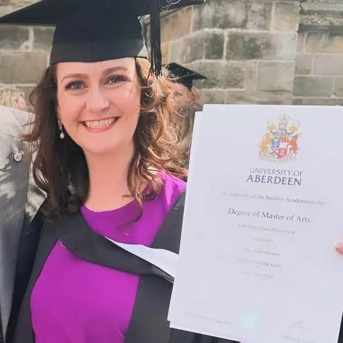 Jenna is dressed in graduation robes and holds up her degree
