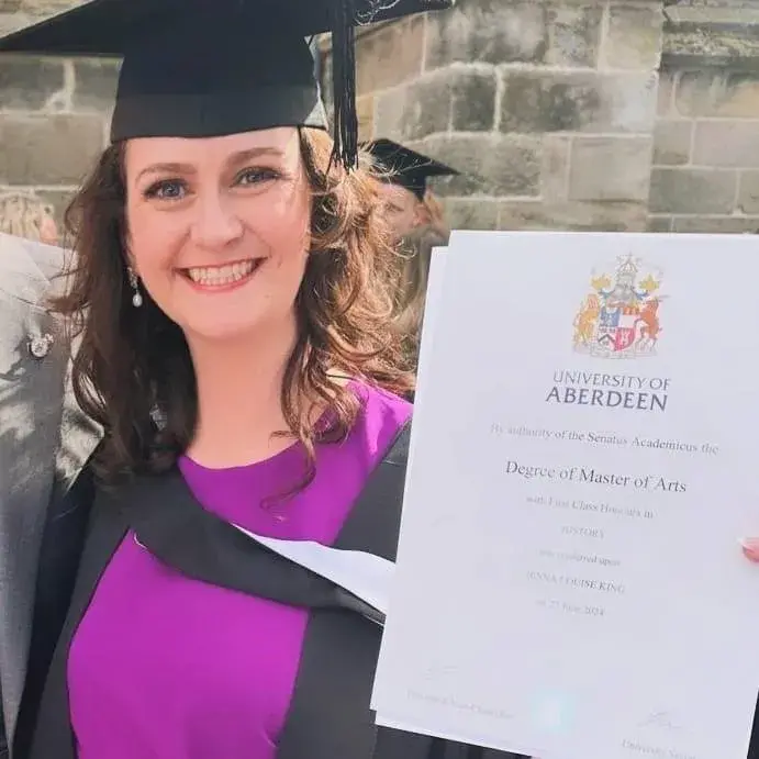 Jenna is dressed in graduation robes and holds up her degree