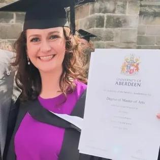 Jenna is dressed in graduation robes and holds up her degree