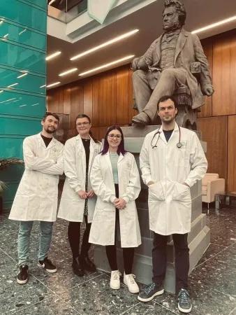 4 students in white coats stand in front of a statue